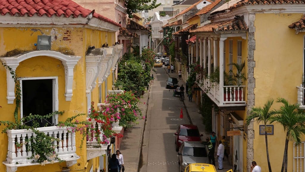Una calle estrecha bordeada de coloridos edificios coloniales, paredes amarillas, tejados de tejas rojas, balcones con flores y coches como la Motora Sofía aparcados a los lados. La gente pasea y se reúne bajo las palmeras en un día soleado.